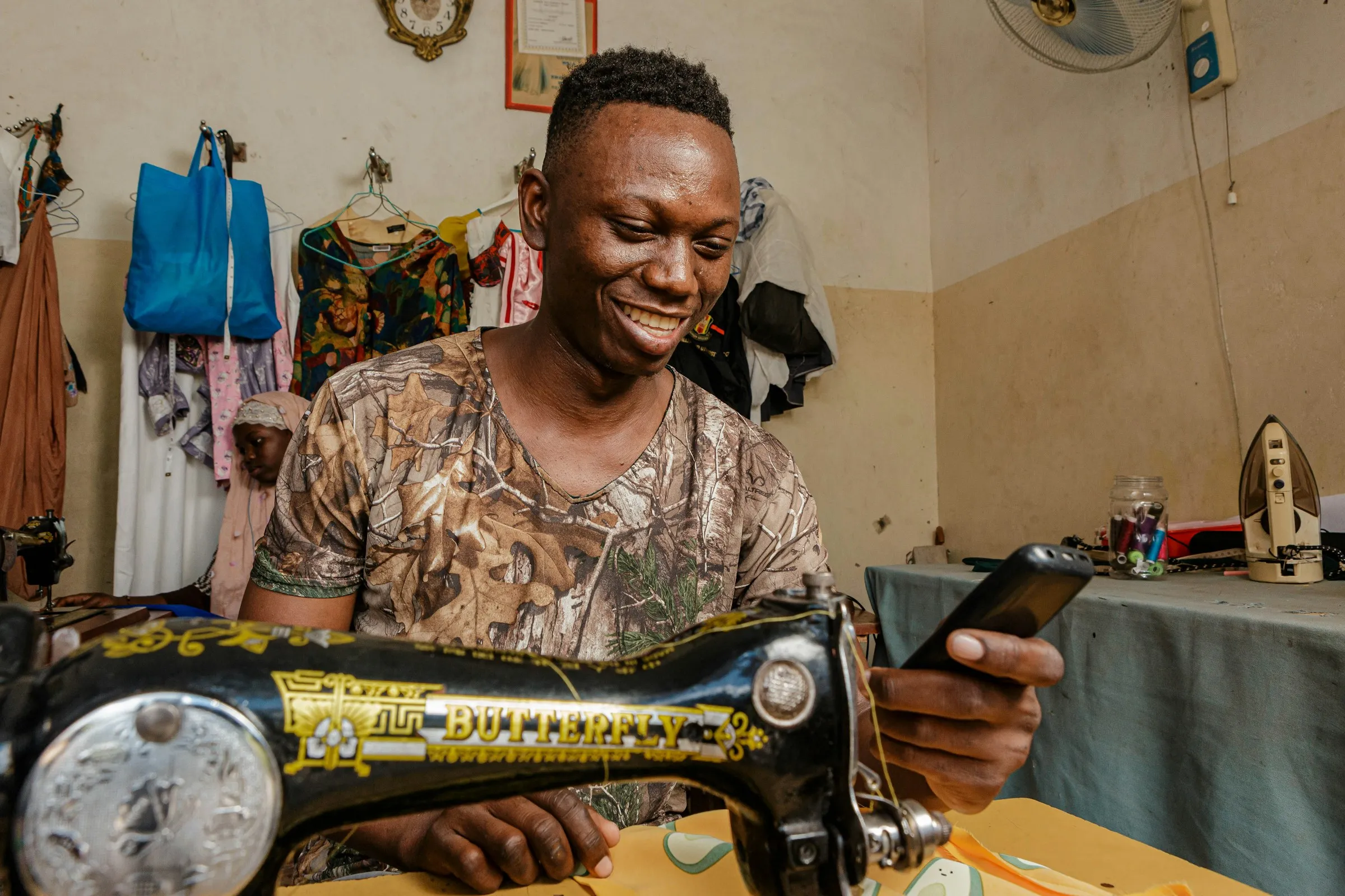 A tailor at his sewing machine in his workshop, checking an order on his phone.
